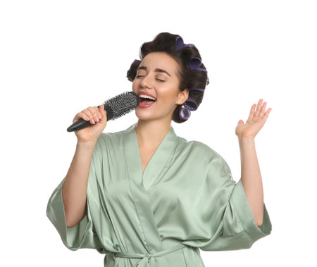 Happy young woman in silk bathrobe with hair curlers singing into hairbrush on white backgroundの写真素材