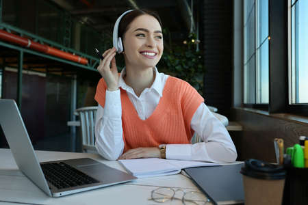 Young female student with laptop and headphones studying at table in cafe