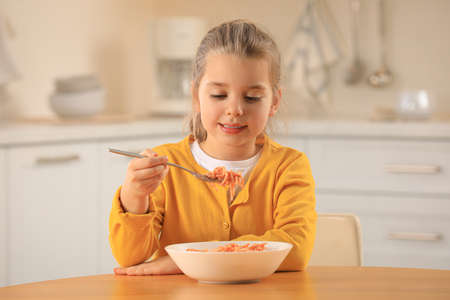 Cute little girl eating tasty pasta at table in kitchenの写真素材