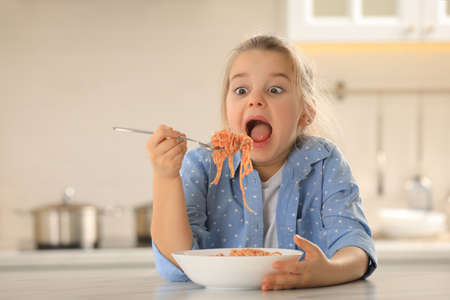 Cute little girl eating tasty pasta at table in kitchenの写真素材