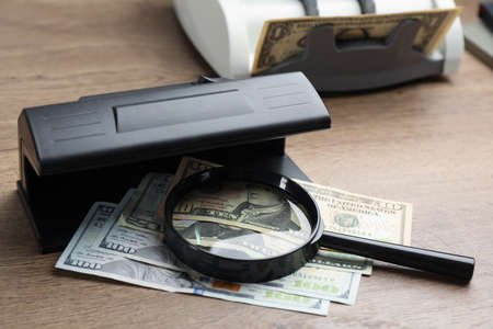Modern currency detector with dollar banknotes and magnifying glass on wooden table. money examination deviceの写真素材