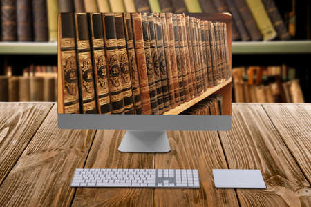 online library. Modern computer on wooden table and shelves with books indoorsの写真素材