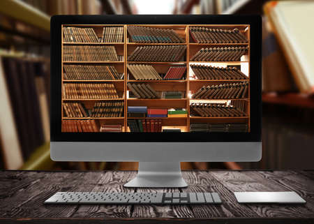 online library. Modern computer on wooden table and shelves with books indoorsの写真素材