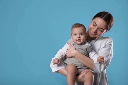Young pediatrician with cute little baby on light blue background. Space for text