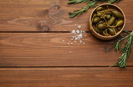 Tasty capers in bowl, salt and rosemary on wooden table, flat lay. Space for textの写真素材