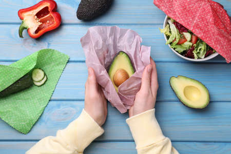 Woman packing half of fresh avocado into beeswax food wrap at light blue wooden table, top viewの写真素材