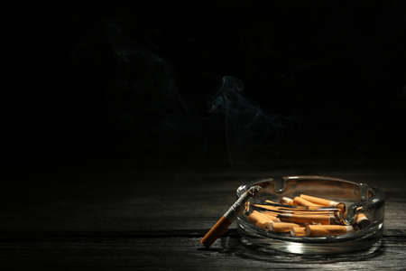 Glass ashtray with stubs and smoldering cigarette on dark wooden table against black background. Space for textの写真素材
