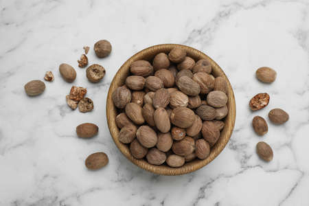 Bowl with nutmeg seeds on white marble table, flat lay