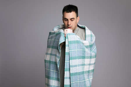 Young man wrapped in blanket with cup of hot drink suffering from cold on gray backgroundの写真素材