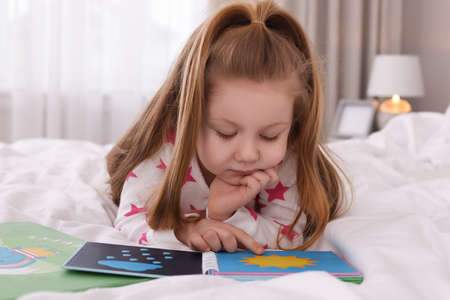 Cute little girl reading book on bed at home