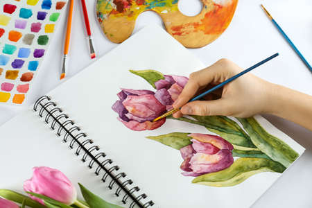 Woman painting tulips in sketchbook and supplies at white table, closeupの写真素材