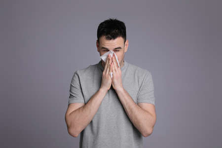 Young man blowing nose in tissue on gray background. cold symptomsの写真素材