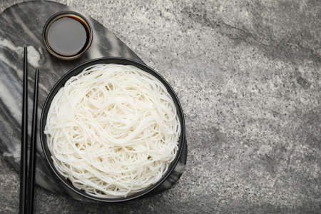 Bowl with cooked rice noodles, soy sauce and chopsticks on gray table, top view. Space for text