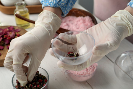 Woman in gloves filling bath bomb mold with flower buds at white table, closeupの写真素材
