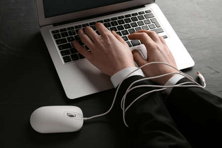 internet addiction. Closeup of man typing on laptop at black table, hands tied with computer mouse cableの写真素材