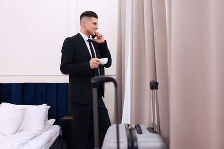 Handsome businessman drinking coffee while talking on phone near window in hotel roomの写真素材