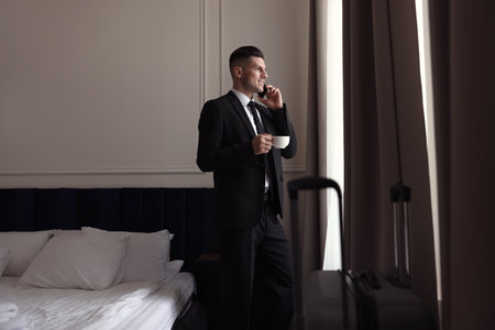 Handsome businessman drinking coffee while talking on phone near window in hotel roomの写真素材