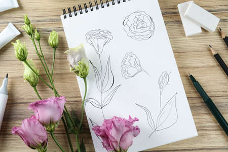 Sketches of flowers in notebook, eustoma and art supplies on wooden table, flat lay