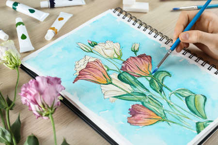 Woman painting eustoma in sketchbook on wooden table, closeupの写真素材