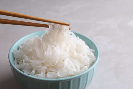 Taking rice noodles with chopsticks from bowl at light gray table, closeup