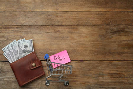 Card with word Cashback, shopping cart and dollar banknotes in wallet on wooden background, flat...