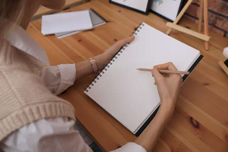 Woman drawing in sketchbook with pencil at wooden table, closeup