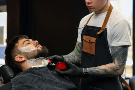 Professional hairdresser applying shaving foam onto client's beard in barbershopの写真素材