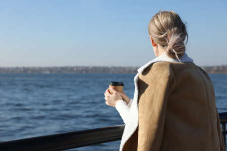 Lonely woman with cup of drink near river on sunny day, back view