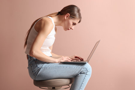 Woman with bad posture using laptop while sitting on stool against pale pink backgroundの写真素材