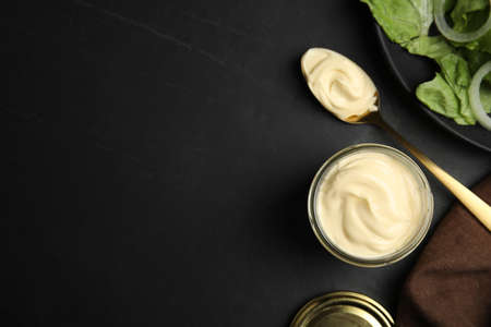 Jar and spoon with delicious mayonnaise near fresh salad on black table, flat lay. Space for textの写真素材