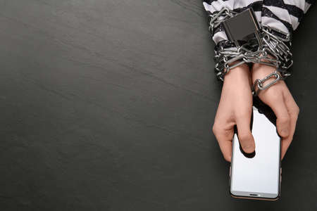Prisoner holding smartphone in chained hands at black table, top view. internet addiction