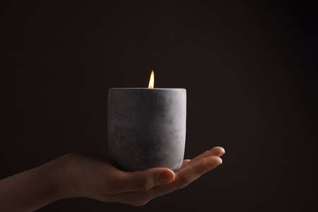 Woman with lit candle in concrete holder against dark brown background, closeupの写真素材