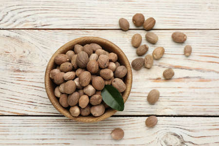 Bowl with nutmeg seeds and green leaf on white wooden table, flat layの写真素材