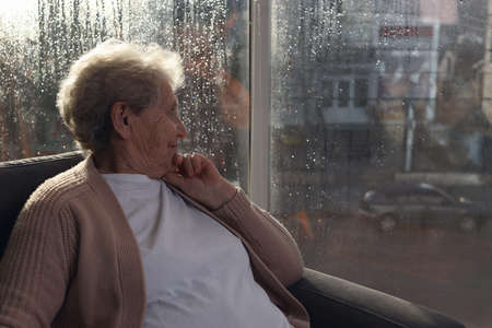 Elderly woman looking out of window on rainy day, space for text. Loneliness conceptの写真素材