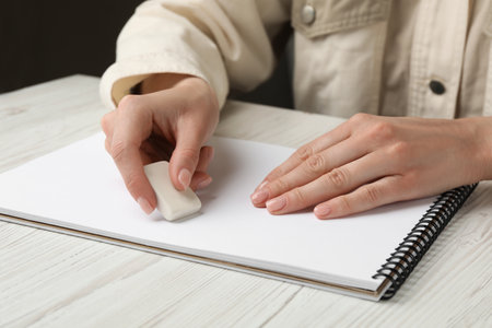 Woman erasing something in notebook at white wooden table, closeup
