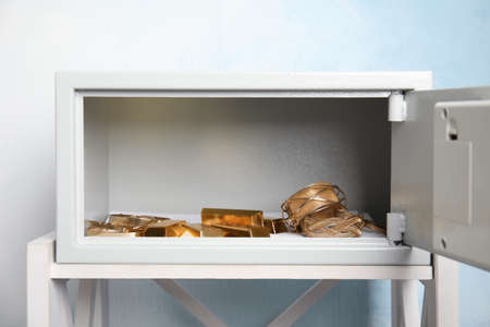 Open steel safe with gold bars and jewelry on white table against light blue backgroundの写真素材