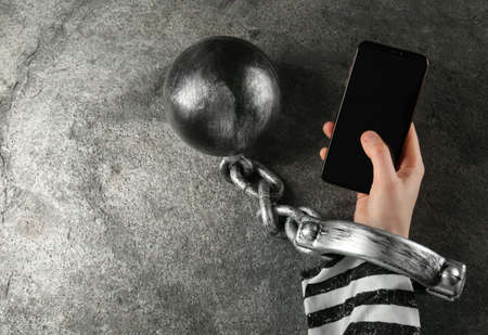 Prisoner shackled with ball and chain holding smartphone at gray table, top viewの写真素材