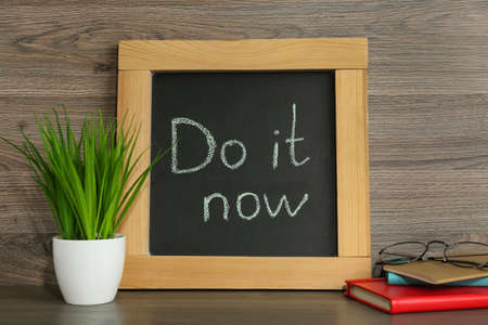 Small chalkboard with motivational quote Do It Now, stationery and plant on wooden table