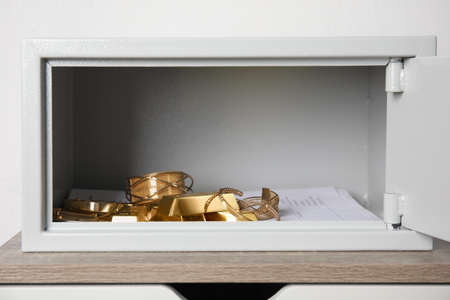 Open steel safe with gold bars and jewelry on wooden table against white backgroundの写真素材