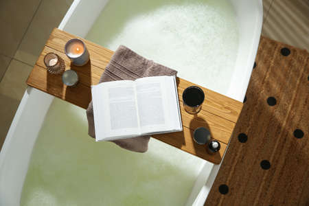 Wooden bath tray with open book, glass of wine and cosmetic products on tub indoors, top viewの写真素材
