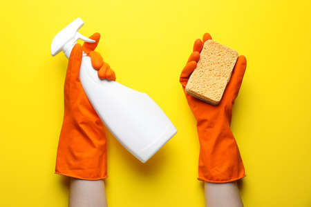 Woman in rubber gloves holding sponge and detergent on yellow background, top view