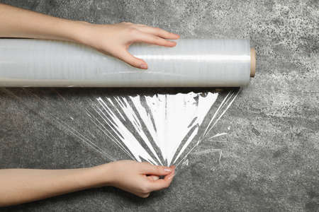 Woman unrolling transparent plastic stretch wrap on gray stone background, top view