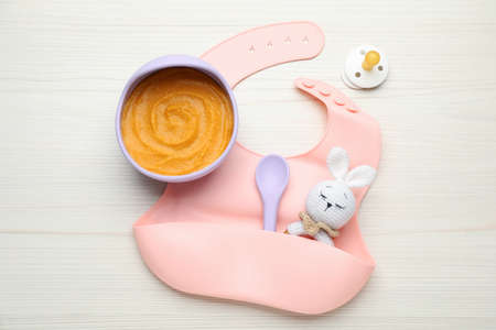 Silicone baby bib, toy and plastic dishware with healthy food on white wooden table, flat lay