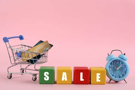 Word Sale made with colorful cubes, alarm clock and credit cards in shopping cart on pink backgroundの写真素材