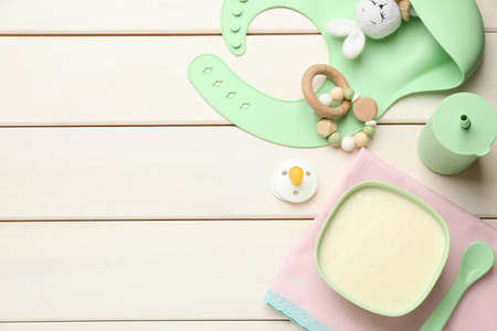 Silicone baby bib, toys and plastic dishware with healthy food on white wooden table, flat lay....