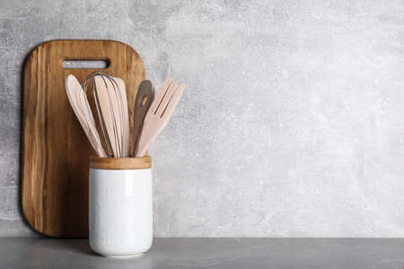 Holder with wooden kitchen utensils and cutting board on gray table. Space for text