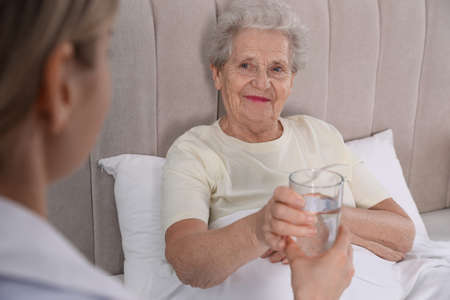 Young caregiver giving water to senior woman in the bedroom. home health care serviceの写真素材
