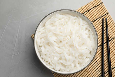 Bowl with cooked rice noodles, straw mat and chopsticks on light grey table, top view