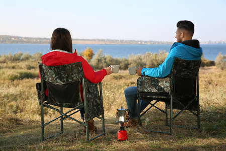 Couple resting in camping chairs and enjoying hot drink outdoors, back viewの写真素材