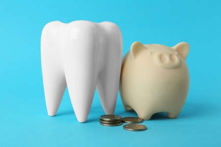 Ceramic model of tooth, piggy bank and coins on light blue background. Expensive treatmentの写真素材